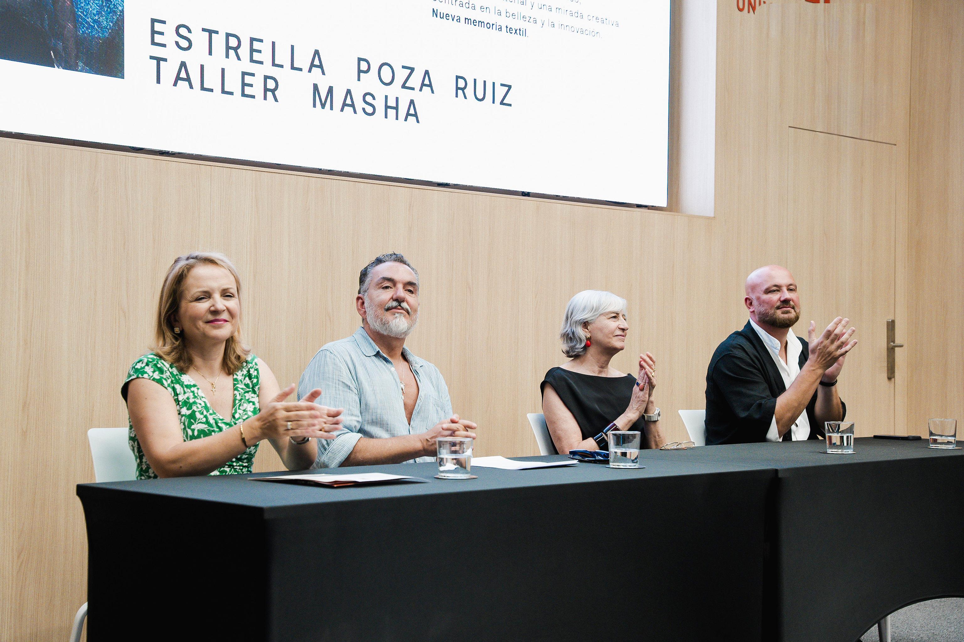 Un grupo de cuatro personas aplaude en un evento, sentados en una mesa al frente de una presentación.