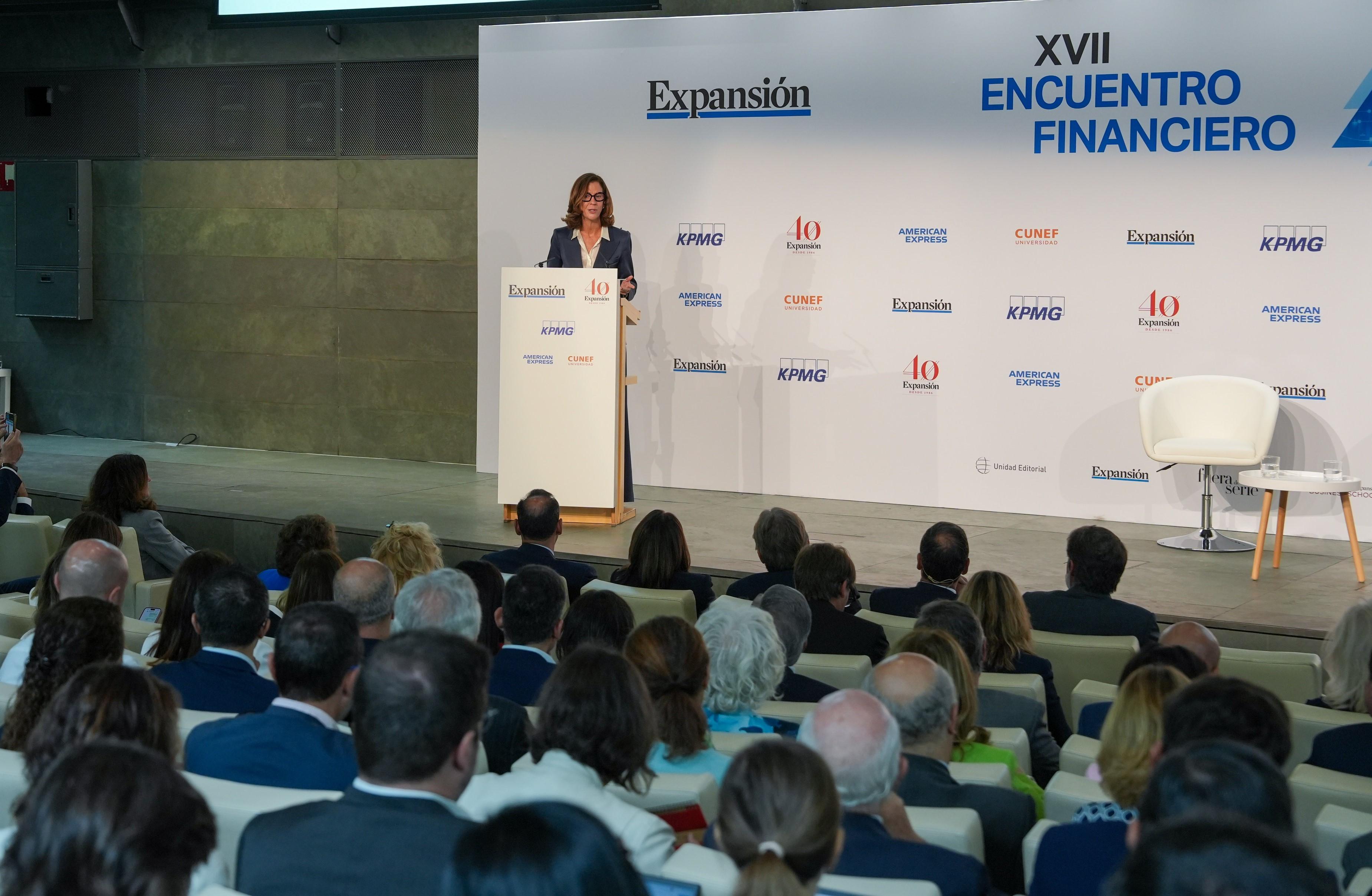 Una ponente en un evento financiero presenta ante una audiencia en un escenario profesional.