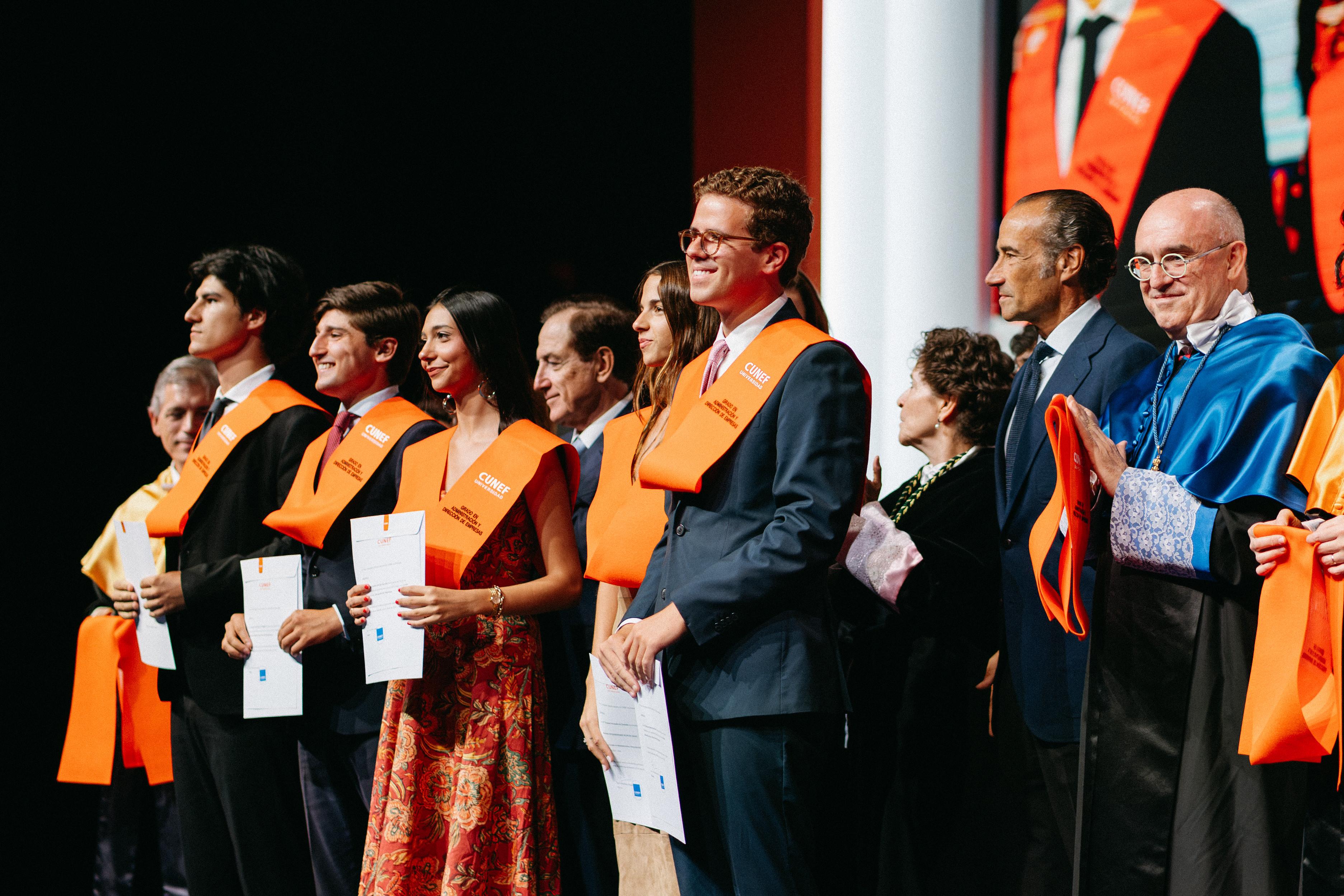 Un grupo de personas en un evento de graduación, posando con diplomas y cintas naranjas.