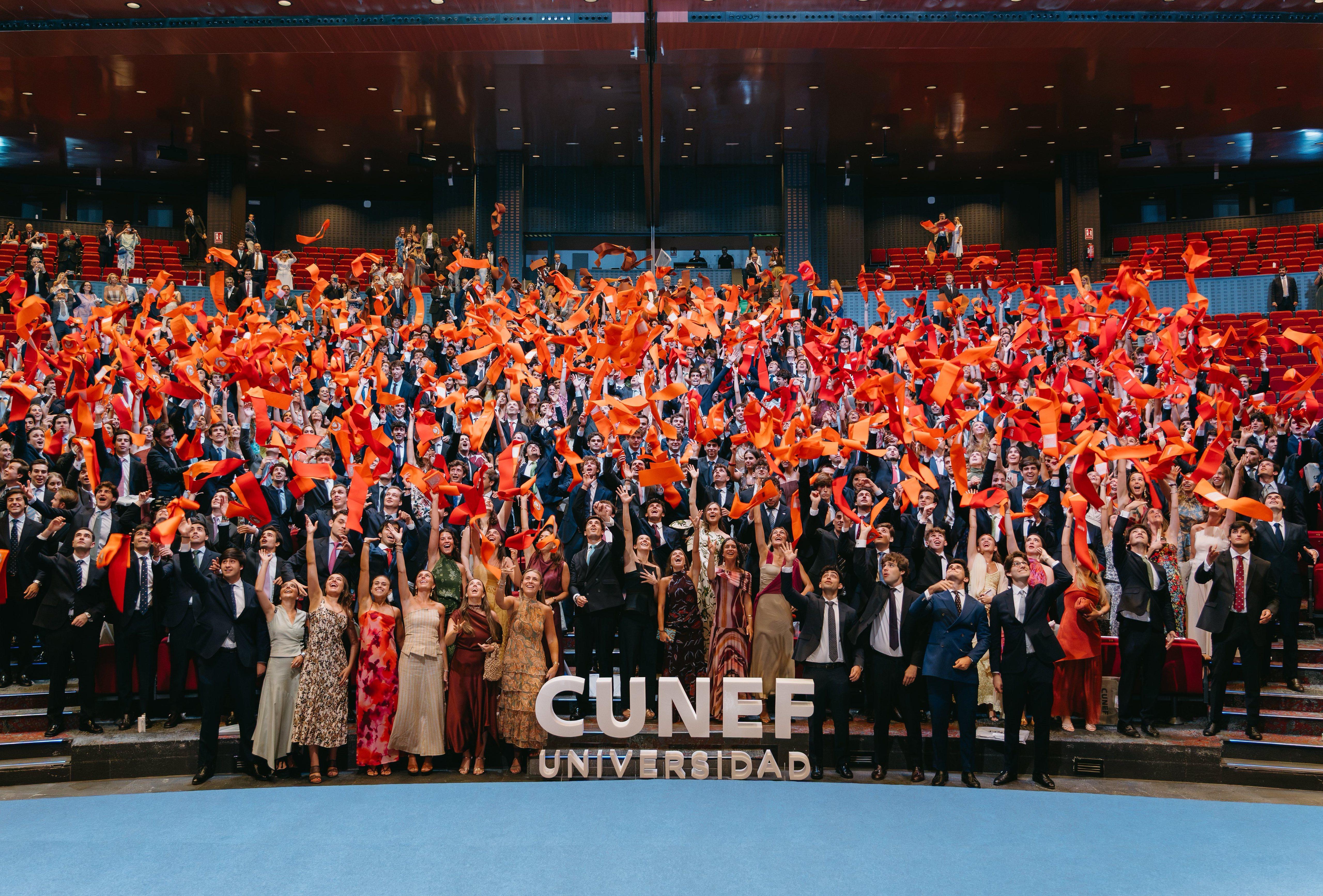 Un gran grupo de graduados celebra su éxito alzando pañuelos naranjas en un auditorio.