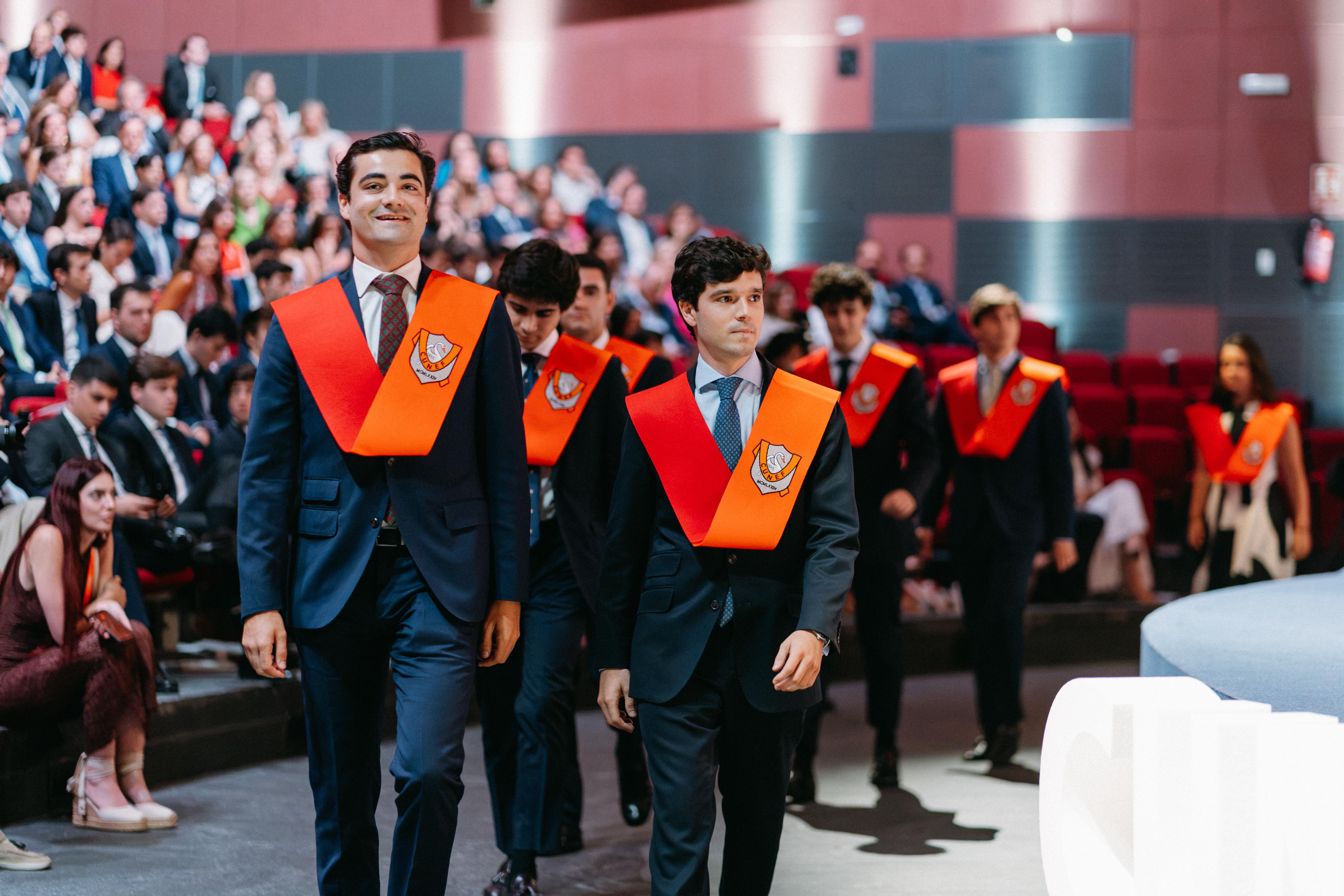 Un grupo de jóvenes con túnicas de graduación camina en un auditorio durante una ceremonia académica.