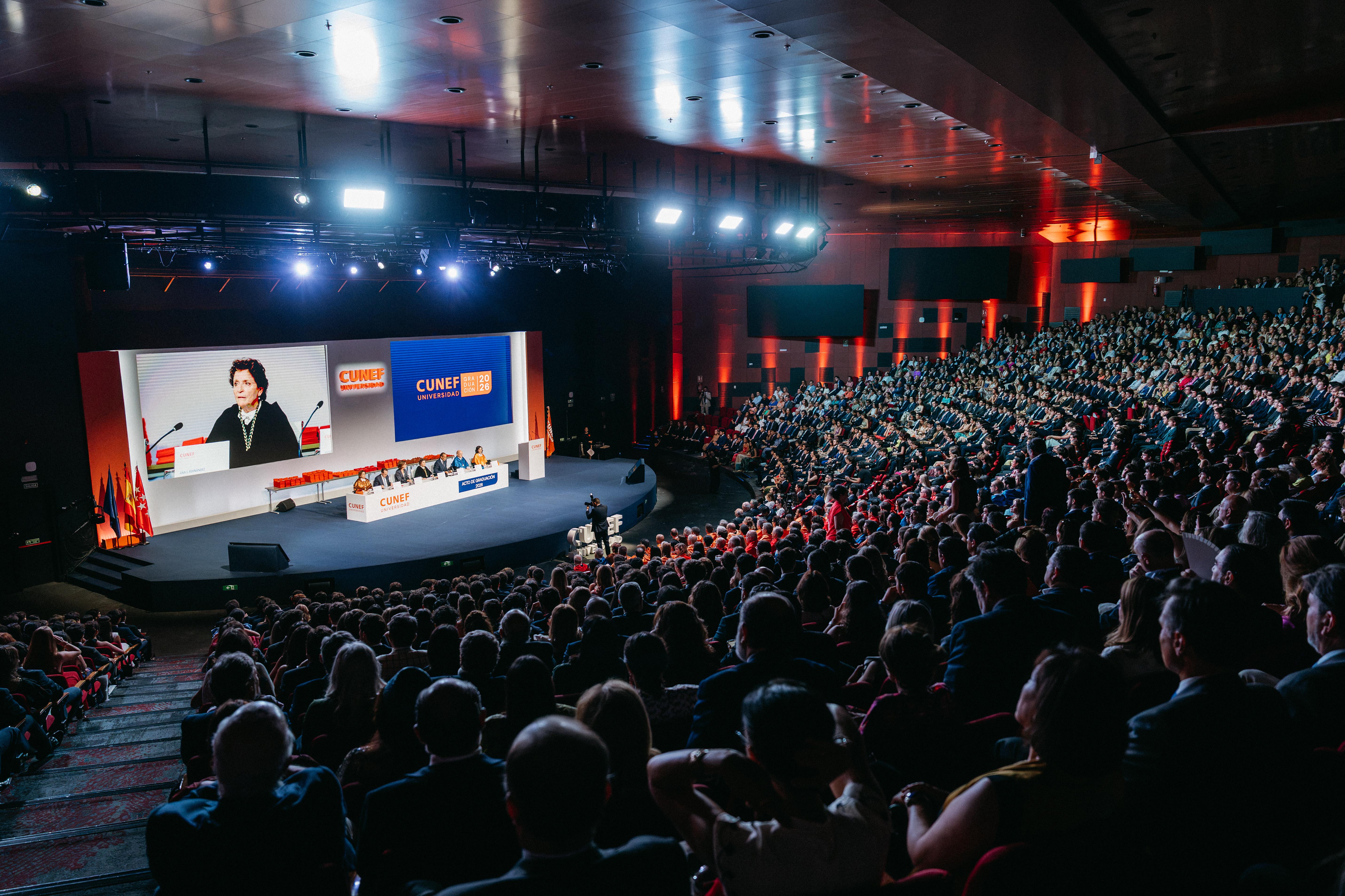 Un auditorio lleno de gente asiste a un evento con una ponente en un escenario iluminado.