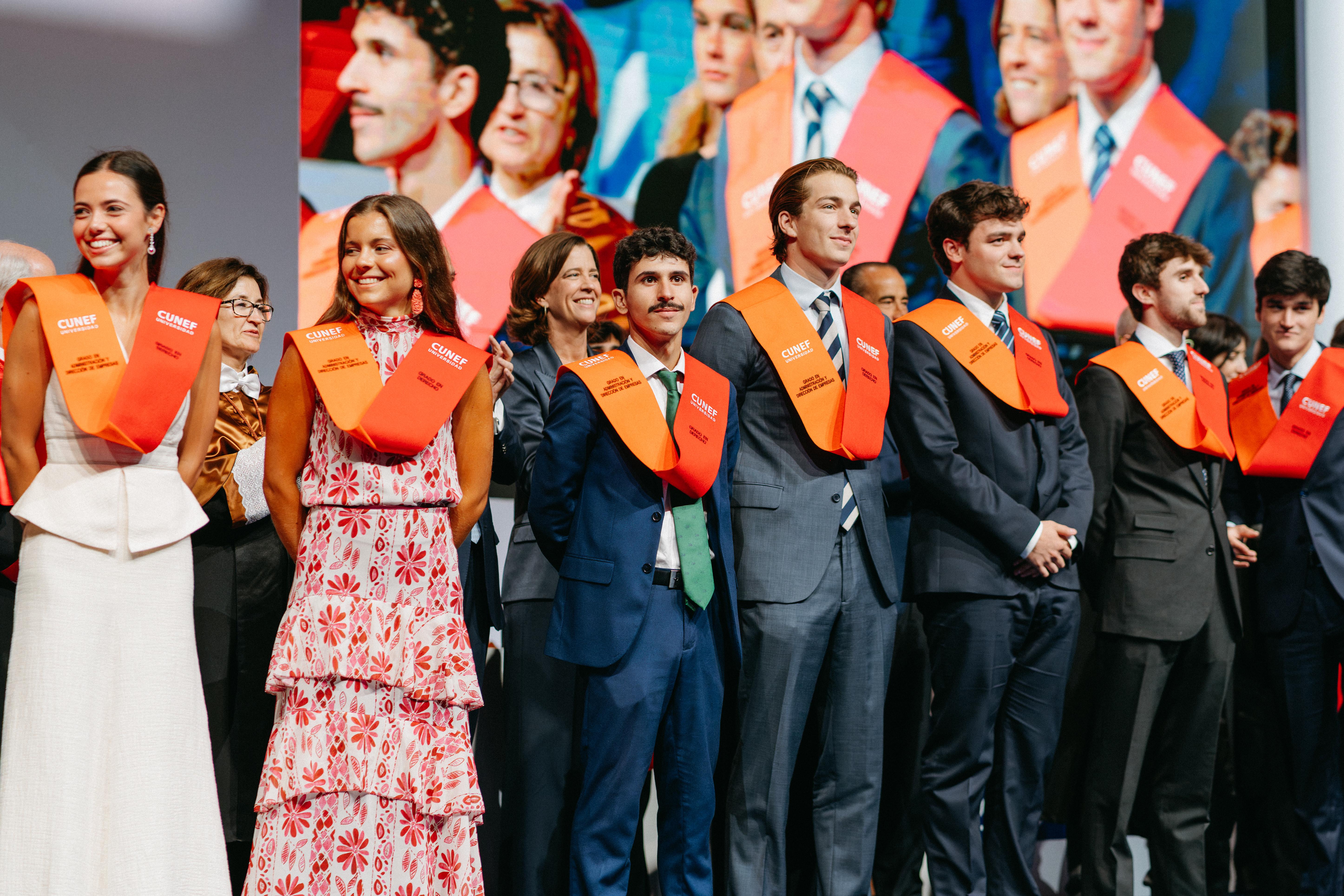 Un grupo de personas en un evento de graduación, vistiendo togas y con bandas naranjas al frente.