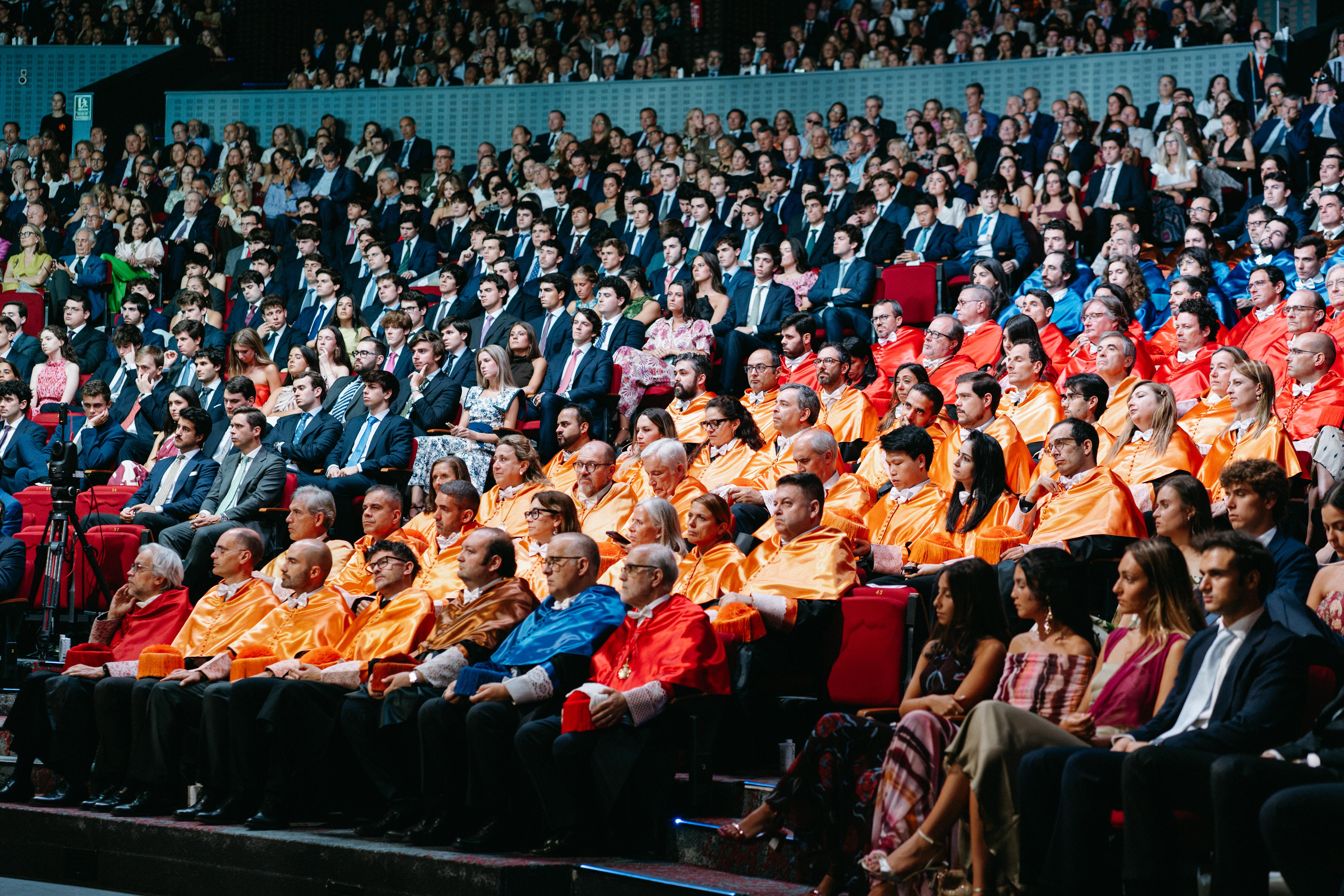 Una multitud de espectadores en un evento, algunos con trajes naranjas y otros con vestimenta formal.