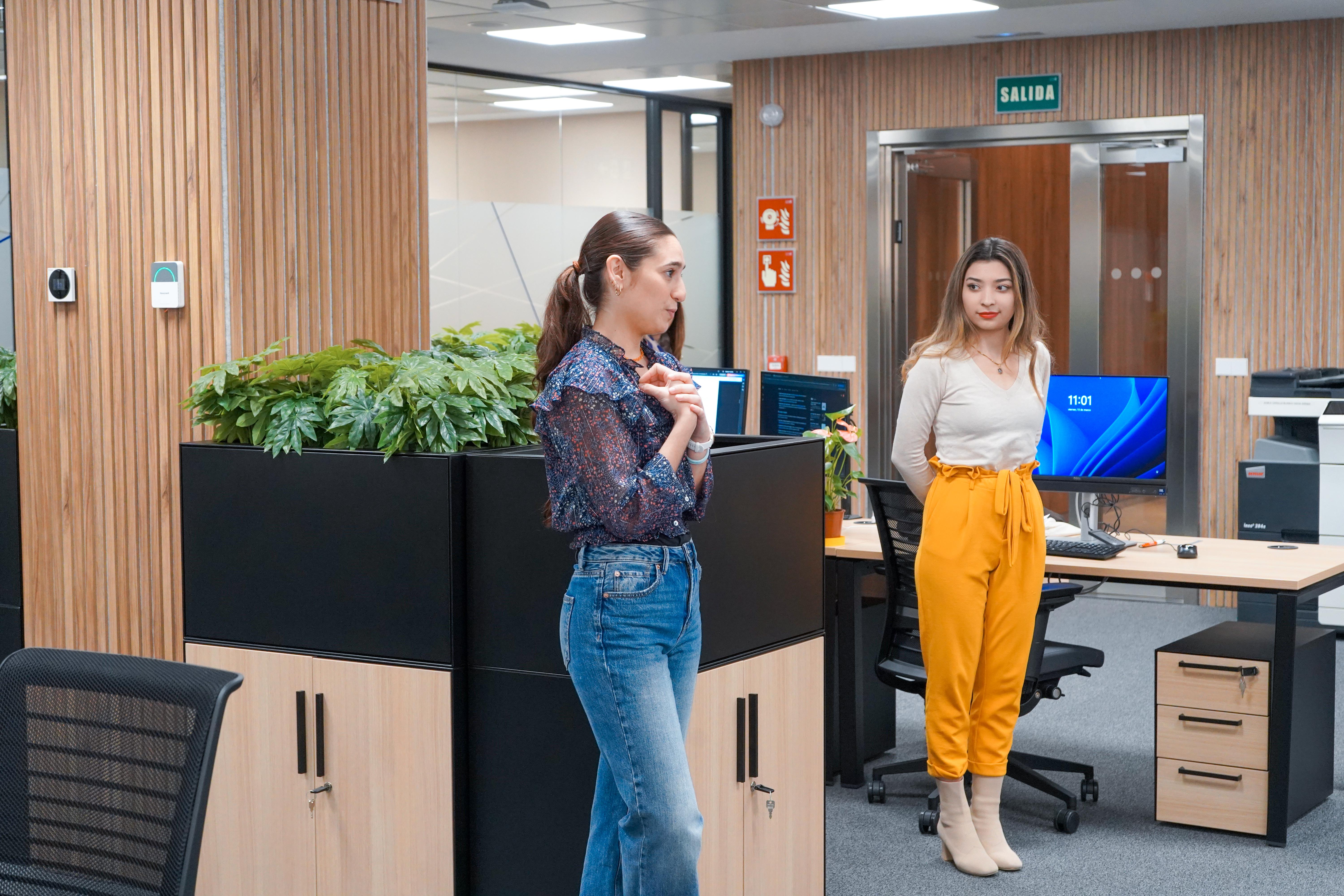 Dos mujeres están en una oficina moderna, una de pie frente a un escritorio y la otra a su lado, conversando en un ambiente profesional.