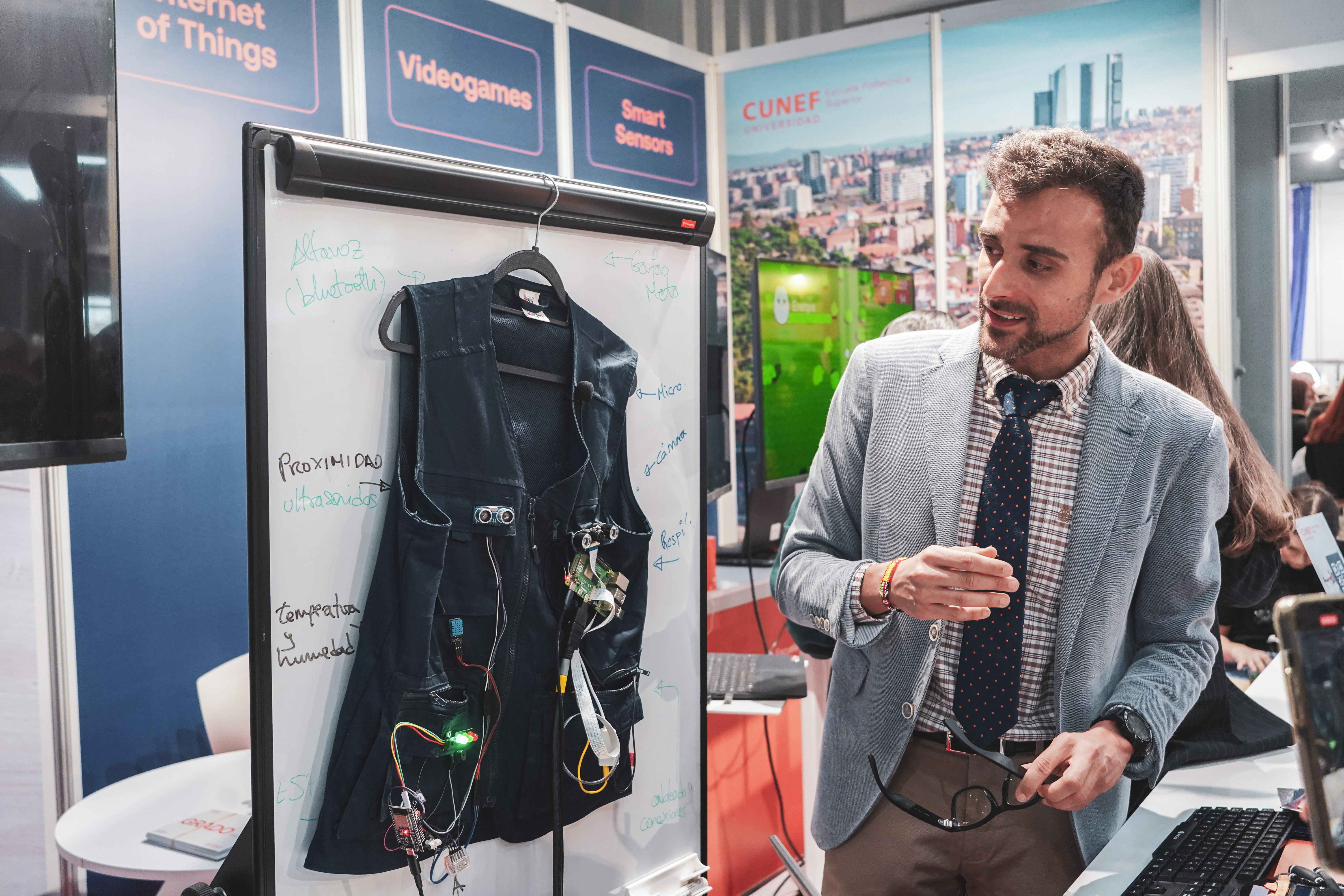 Un hombre presenta un chaleco tecnológico en un evento, explicando sus características frente a un panel de información.