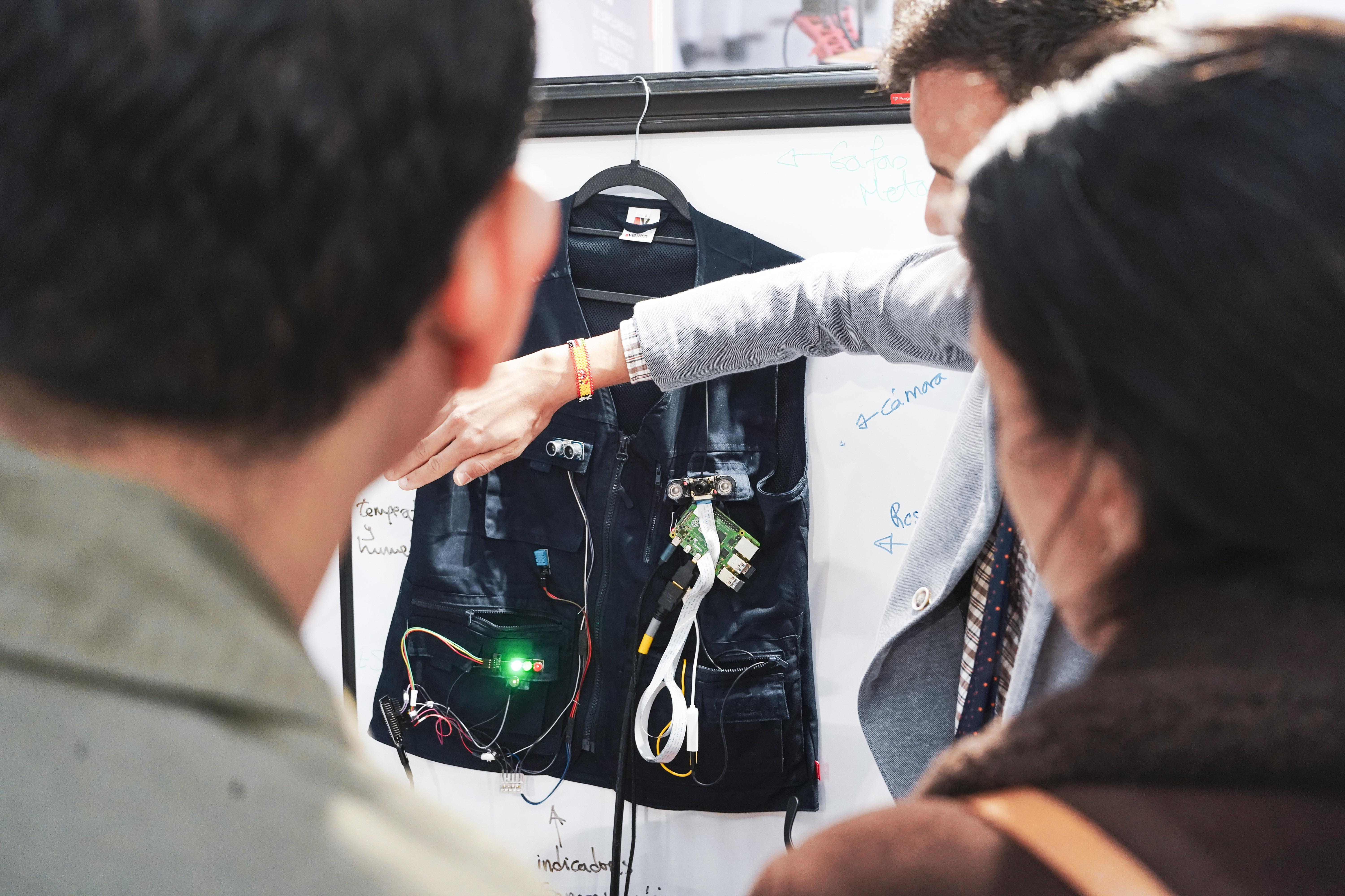 Una persona explica un chaleco tecnológico con componentes electrónicos a dos oyentes interesados.