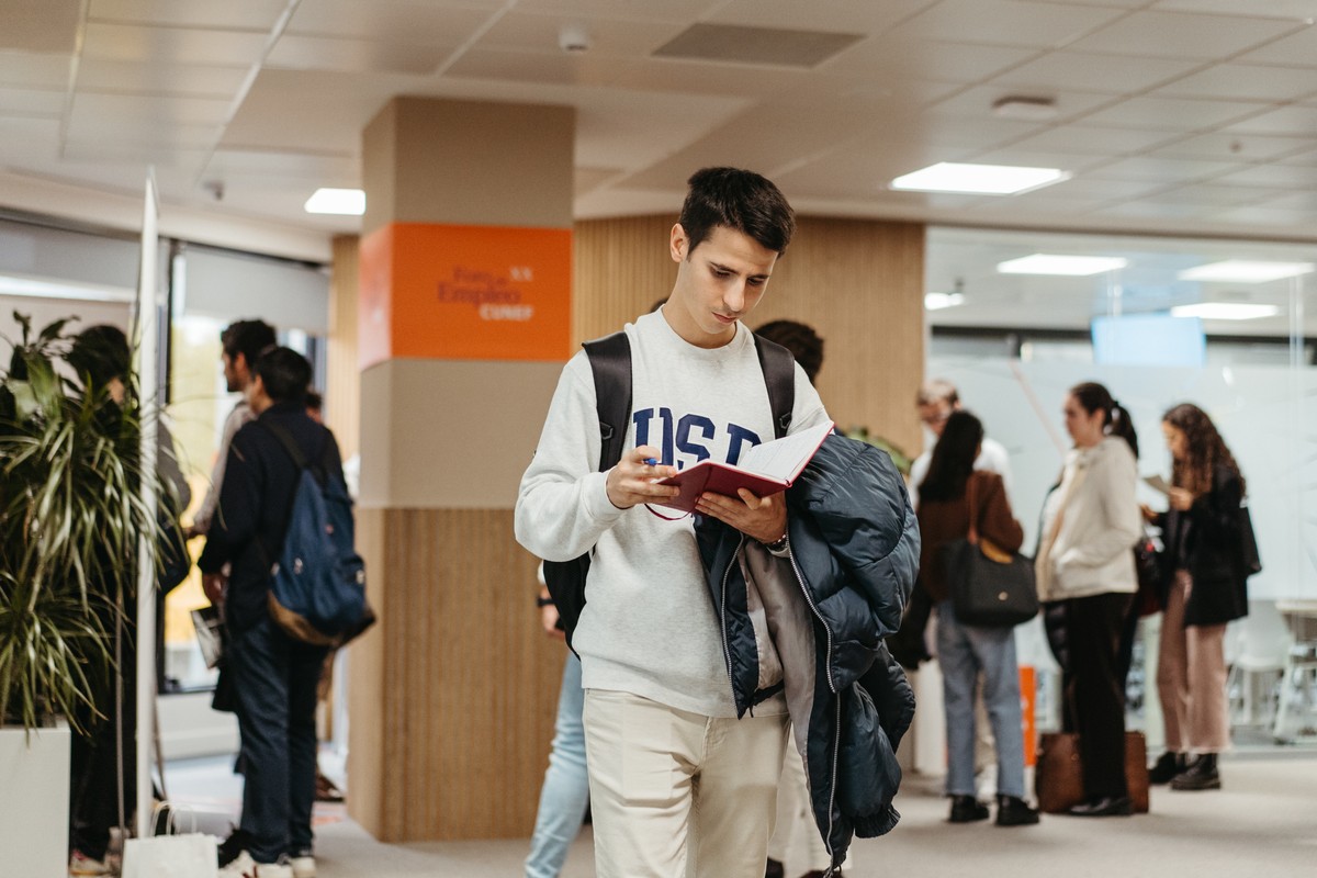 Un estudiante de pie en un ambiente académico, leyendo un libro mientras otros caminan a su alrededor.