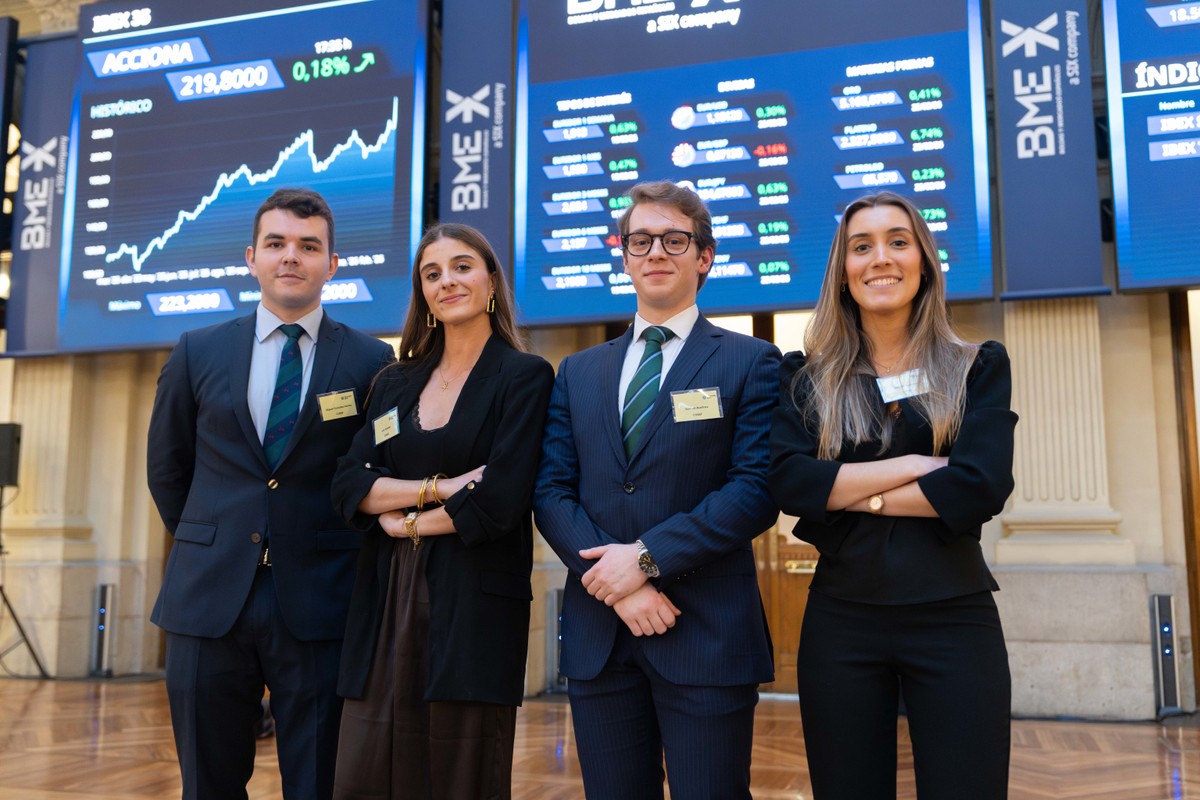 Cuatro jóvenes profesionales posan en un entorno financiero con pantallas que muestran datos del mercado.
