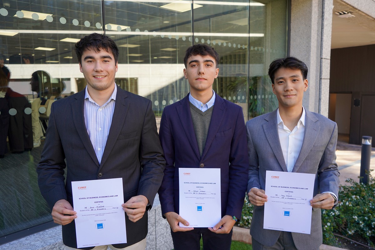 Tres jóvenes posan sonrientes con certificados en manos frente a un edificio moderno.