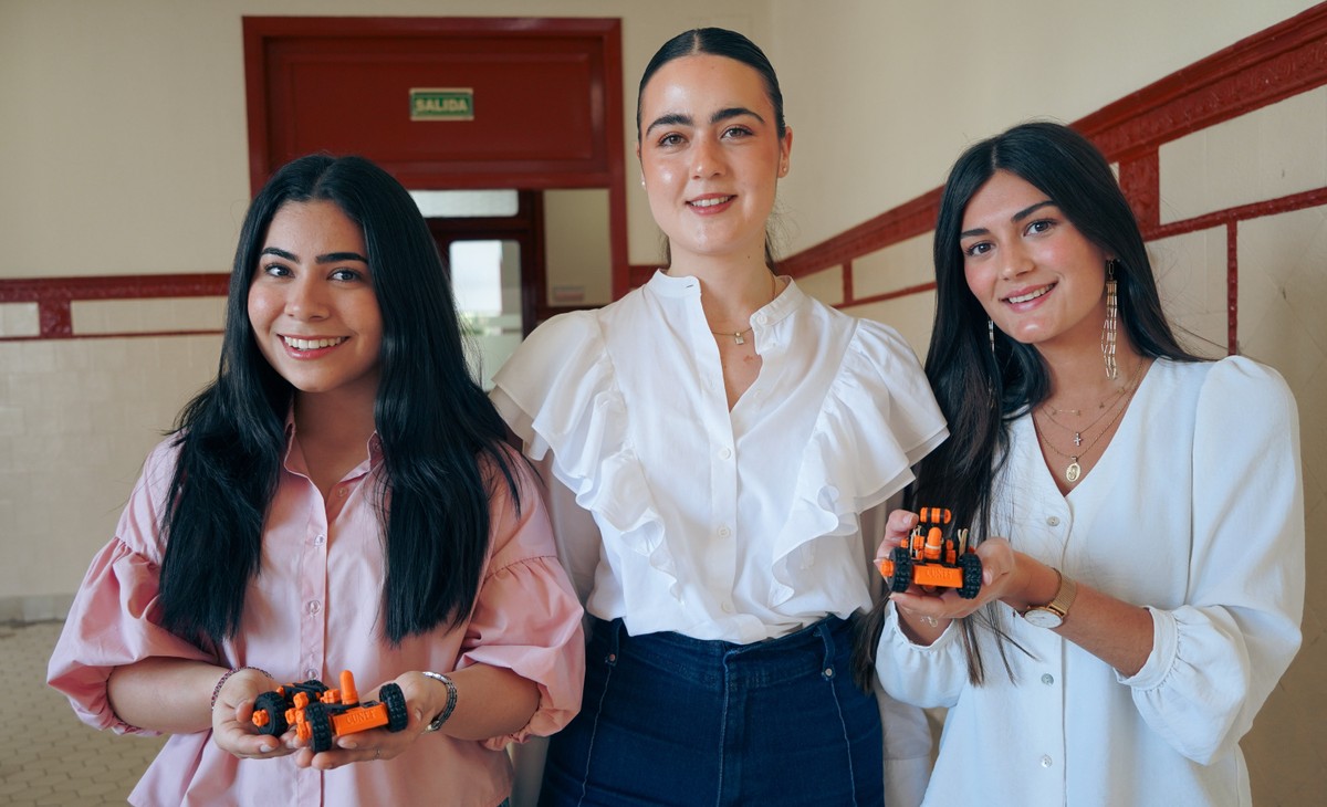 Tres mujeres sonrientes sostienen pequeños objetos en sus manos en un pasillo luminoso.