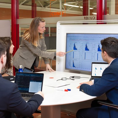 Una mujer presenta datos a un grupo de personas en una reunión alrededor de una mesa con computadoras portátiles.