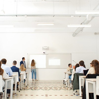 Una sala de clases con estudiantes sentados al frente y dos personas en la pizarra.