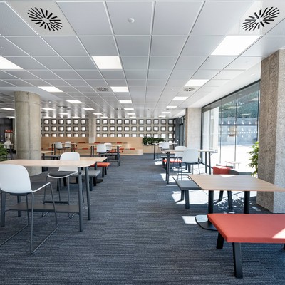 Espacio interior moderno con mesas y sillas, rodeado de ventanas que permiten la entrada de luz natural.