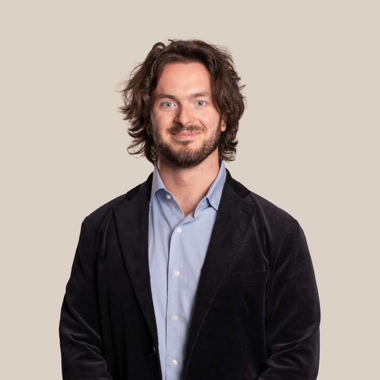 Un hombre sonriente vestido con un blazer oscuro y una camisa clara, posa con las manos juntas frente a un fondo sencillo.