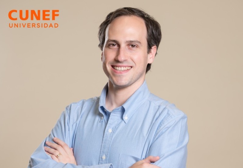 Un hombre sonriente con una camisa azul claro posa con los brazos cruzados frente a un fondo beige.